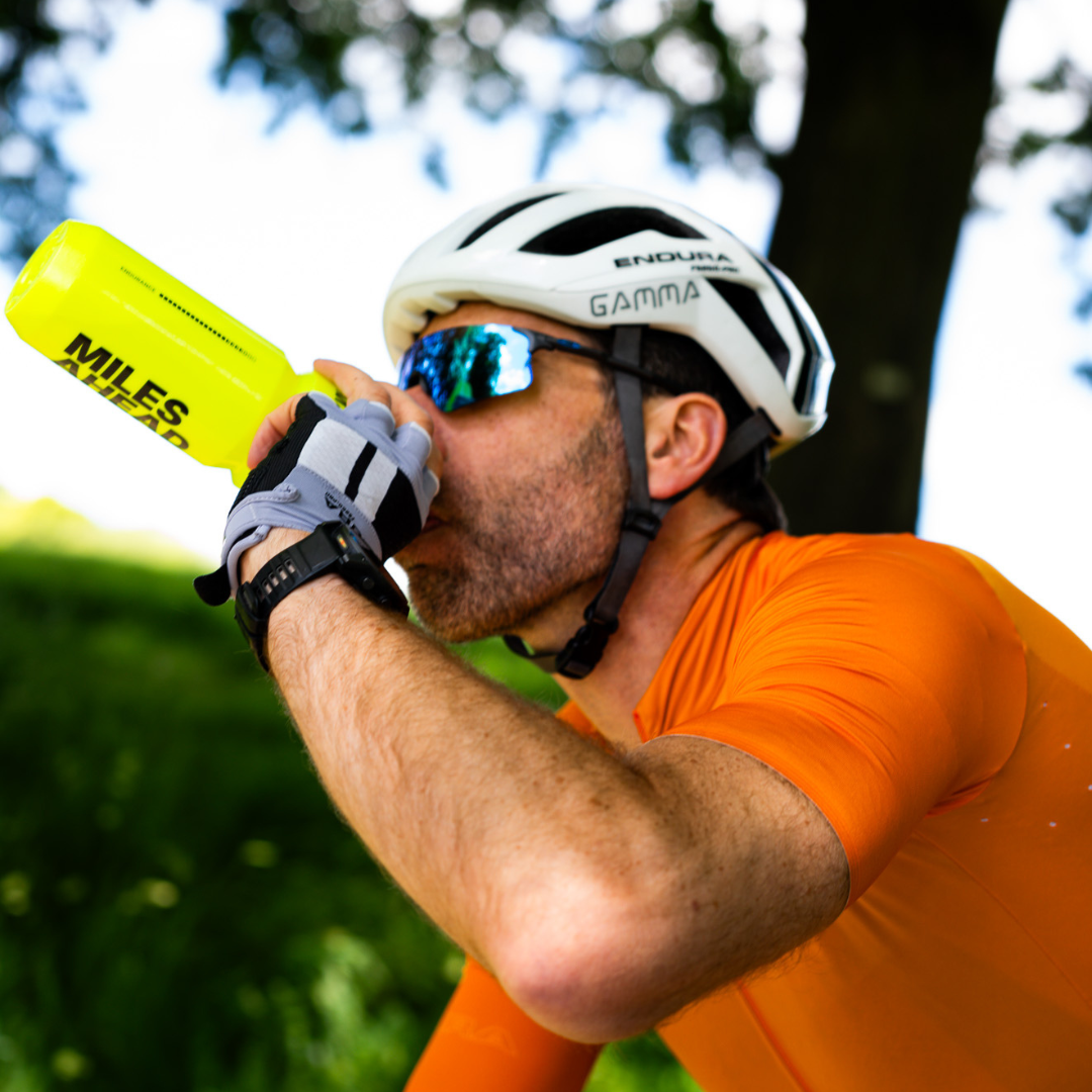 A cyclist wearing a white Enigma helmet and sunglasses drinks from a bright yellow MOVE Pre-Workout bottle labeled 'MILES AHEAD' in an outdoor setting.